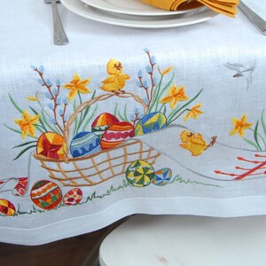 May include: White linen tablecloth with embroidered Easter design. The design features a basket filled with colorful eggs, yellow chicks, and daffodils.