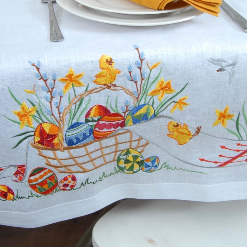 May include: White linen tablecloth with embroidered Easter design. The design features a basket filled with colorful eggs, yellow chicks, and daffodils.