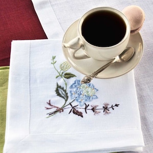 Könnte beinhalten: Weiße Leinen-Serviette mit gesticktem blauem und grünem Blumenmuster. Eine Tasse Kaffee und eine Untertasse liegen auf der Serviette.