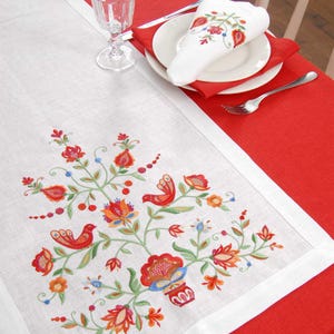 Peut inclure: Une nappe en lin blanc avec une broderie florale détaillée en rouge, orange, bleu et vert. Le chemin de table est posé sur une nappe rouge, avec un ensemble de serviettes et d'assiettes assorties.