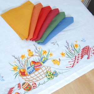 May include: A white linen tablecloth with an embroidered Easter design featuring a basket of colorful eggs, chicks, and daffodils. The tablecloth is decorated with a border of red and white embroidered flowers. Seven colorful napkins are folded and arranged on the tablecloth.