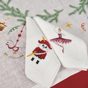 May include: White cloth napkins with embroidered Christmas designs. One napkin features a nutcracker soldier in red and gold, the other a ballerina in a burgundy dress. The background has a Christmas tree and toy designs.