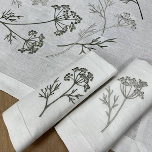以下が含まれることがあります： 白のリネンナプキンと、グレーとグリーンの刺繍されたディルの花の小枝が特徴のテーブルクロス。 ナプキンは折りたたまれ、テーブルクロスの上に置かれ、コーディネートされたテーブルセッティングを作成します。