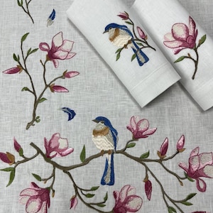 Op de afbeelding: Wit linnen met geborduurde blauwborstjes en roze magnoliabloemen. Twee opgerolde witte servetten met bijpassende vogel- en bloemborduurwerk staan rechtsboven. Het ontwerp heeft een natuurthema.