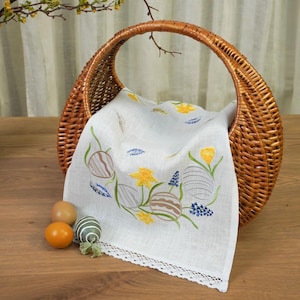 May include: A woven wicker basket holds a white linen cloth embroidered with Easter egg and floral designs in yellow, blue, and green. Three eggs in shades of brown, orange, and green sit in front of the basket on a wooden surface. A sprig of a plant is in the background.