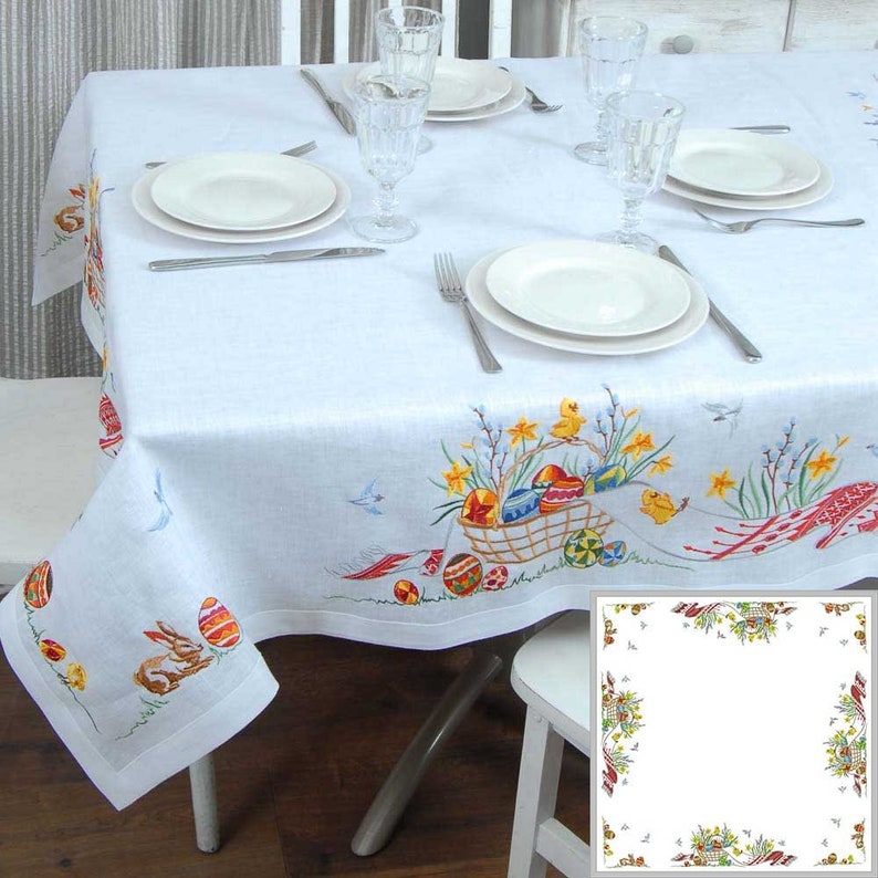 May include: White linen tablecloth with an embroidered Easter design. The design features a basket of colorful eggs, chicks, and spring flowers. The tablecloth has a scalloped edge.