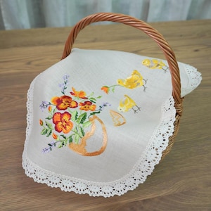 Op de afbeelding: Een geweven rieten mand met een handvat, bedekt met een witte doek geborduurd met oranje en gele bloemen in een vaas, en gele kuikens. De doek heeft een witte kanten rand.