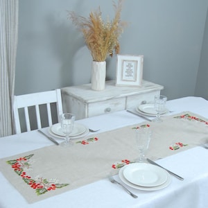 May include: A beige linen table runner with red berry embroidery. The runner is laid on a white tablecloth and features a simple design of red berries and green leaves. The runner is set for a meal with two place settings, each with a white plate, a glass, and cutlery.