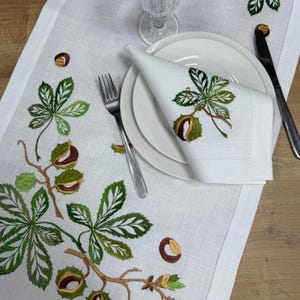 Könnte beinhalten: Weißer Tischläufer aus Leinen mit gestickten grünen Blättern und braunen Kastanien. Eine weiße Serviette mit passender Stickerei liegt auf einem weißen Teller. Eine Gabel und ein Messer sind auf beiden Seiten des Tellers platziert.