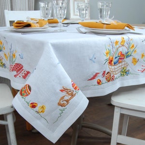 May include: White linen tablecloth with embroidered Easter designs, including a basket of eggs, a bunny rabbit, and spring flowers. The tablecloth is set with two place settings, each with a yellow napkin and a glass of water.