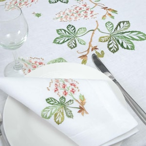 Puede incluir: Juego de mantel y servilleta de lino blanco con diseño floral y de hojas bordadas. El bordado presenta hojas verdes y racimos de flores rosas y rojas. También se ven una copa de vino, un plato y cubiertos.