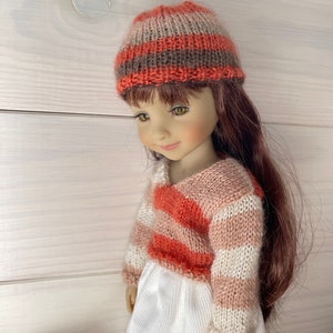 May include: A doll dressed in a knitted hat and sweater. The hat has horizontal stripes in orange, brown, and beige. The sweater is striped in orange, white, and pink, with a V-neck. The doll is wearing a white dress.