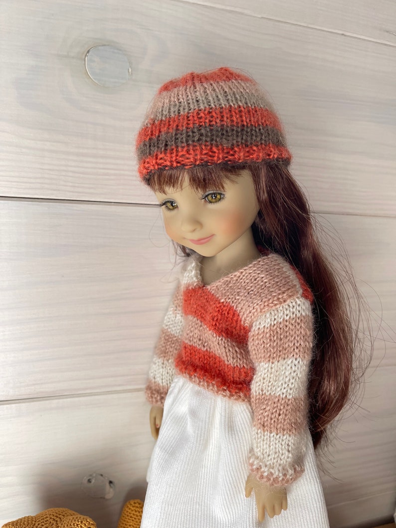 May include: A doll dressed in a knitted hat and sweater. The hat has horizontal stripes in orange, brown, and beige. The sweater is striped in orange, white, and pink, with a V-neck. The doll is wearing a white dress.