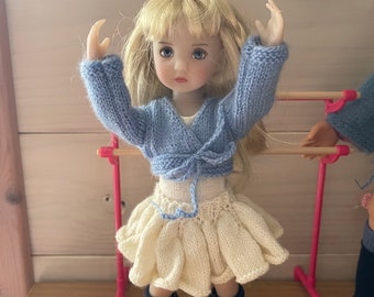May include: A doll wearing a light blue knitted cardigan, a cream knitted skirt, black socks, and white sneakers. The doll has blonde hair and blue eyes.