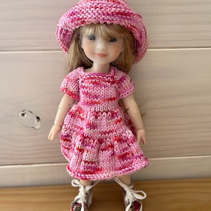 May include: A pink and red striped knitted dress with short sleeves and a matching pink and red striped knitted hat. The doll is wearing white sneakers with floral accents.