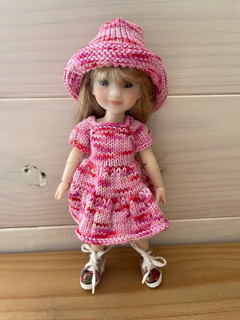 May include: A pink and red striped knitted dress with short sleeves and a matching pink and red striped knitted hat. The doll is wearing white sneakers with floral accents.