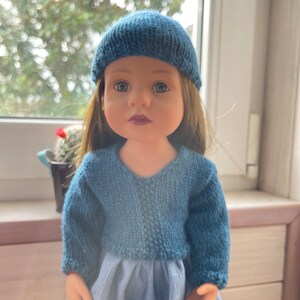 May include: A doll dressed in a knitted blue hat and sweater. The doll has long brown hair and a blue dress. The background is blurred, with a window and a small potted plant in view.