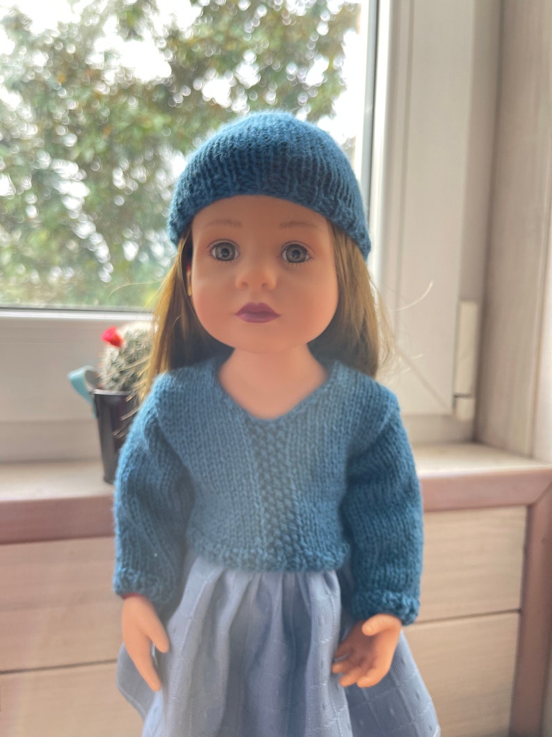 May include: A doll dressed in a knitted blue hat and sweater. The doll has long brown hair and a blue dress. The background is blurred, with a window and a small potted plant in view.