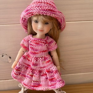 May include: A pink knitted dress and hat set for a doll. The dress has a striped pattern and the hat is a bucket style.