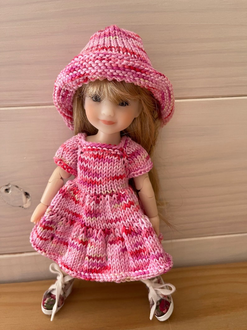 May include: A pink knitted dress and hat set for a doll. The dress has a striped pattern and the hat is a bucket style.