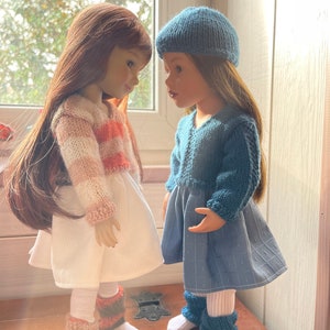May include: Two dolls are shown wearing knitted outfits. One doll has a white dress with a striped pink and cream sweater, and leg warmers. The other doll is in a blue dress, beret, and leg warmers. Both dolls have long hair.