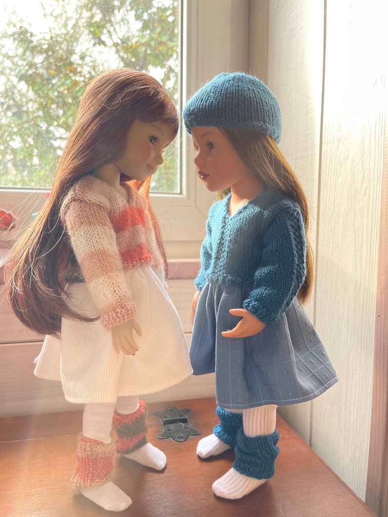May include: Two dolls are shown wearing knitted outfits. One doll has a white dress with a striped pink and cream sweater, and leg warmers. The other doll is in a blue dress, beret, and leg warmers. Both dolls have long hair.