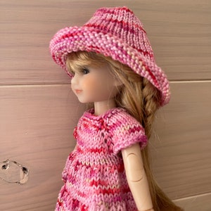 May include: A pink and white striped knitted dress and hat set for a doll. The dress has short sleeves and a round neckline. The hat is a wide-brimmed sun hat.