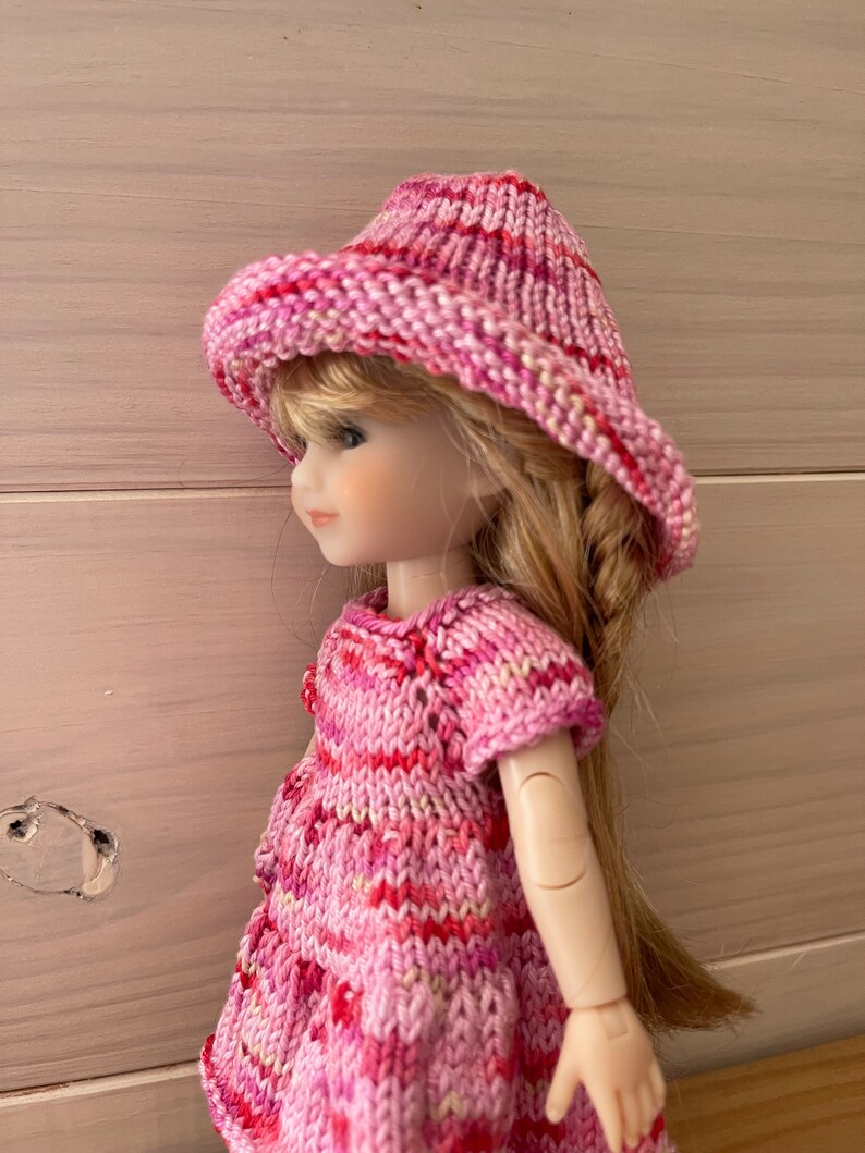 May include: A pink and white striped knitted dress and hat set for a doll. The dress has short sleeves and a round neckline. The hat is a wide-brimmed sun hat.