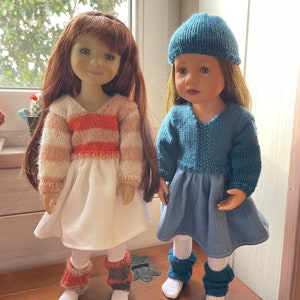 Op de afbeelding: Twee poppen gekleed in handgemaakte gebreide outfits. De ene pop draagt een witte rok, een gestreepte oranje en witte trui en beenwarmers. De andere pop draagt een blauwe jurk, een bijpassende hoed en blauwe beenwarmers. Beide poppen hebben lang haar.
