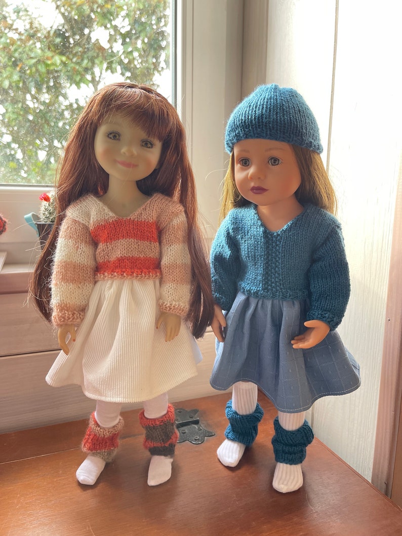 May include: Two dolls are dressed in handmade knitted outfits. One doll wears a white skirt, a striped orange and white sweater, and leg warmers. The other doll is wearing a blue dress, a matching hat, and blue leg warmers. Both dolls have long hair.