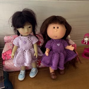 Peut inclure: Deux poupées vêtues de robes lavande faites à la main avec des détails de boutons. L'une a des cheveux foncés en tresses et des baskets bleues. L'autre a des cheveux bruns et des chaussures marron. Les poupées sont assises sur un coussin à motifs floraux roses.