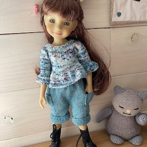 Peut inclure: Une poupée aux cheveux bruns portant un pull bleu et blanc à motifs, un short en jean bleu et des bottes noires. La poupée est debout à côté d'un chat en crochet gris.