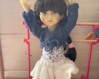 May include: A doll wearing a blue knitted sweater, a white skirt with a floral pattern, and blue sneakers. The doll has long brown hair and is holding her arms up in the air.