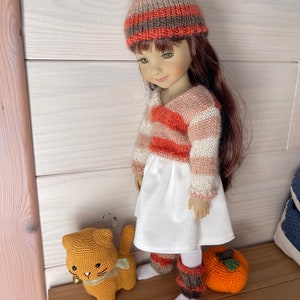 May include: A doll dressed in a knitted hat and sweater with orange, white, and brown stripes, a white dress, and leg warmers. An orange crocheted cat toy with a green ribbon and a crocheted orange pumpkin are also visible.