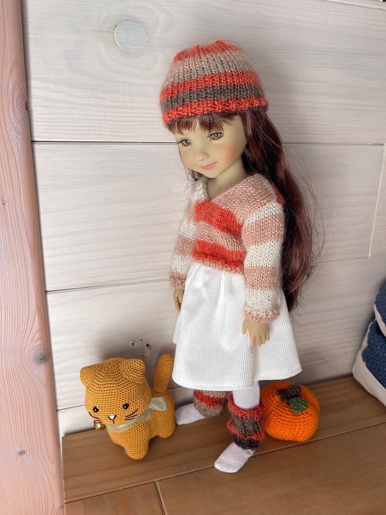 May include: A doll dressed in a knitted hat and sweater with orange, white, and brown stripes, a white dress, and leg warmers. An orange crocheted cat toy with a green ribbon and a crocheted orange pumpkin are also visible.