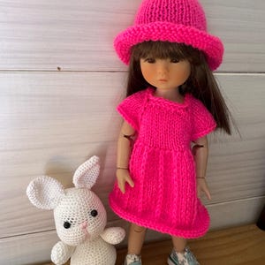 Peut inclure: Une robe et un chapeau en tricot rose pour une poupée, avec un lapin en crochet blanc. La poupée porte des baskets blanches avec des accents verts.