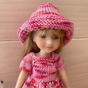 May include: A pink knitted hat and dress set for a doll. The hat is a wide-brimmed style with a pink and white striped pattern. The dress is a short-sleeved, A-line style with a pink and white striped pattern.
