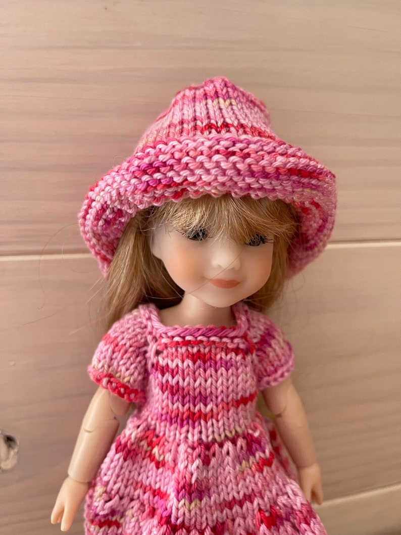 May include: A pink knitted hat and dress set for a doll. The hat is a wide-brimmed style with a pink and white striped pattern. The dress is a short-sleeved, A-line style with a pink and white striped pattern.