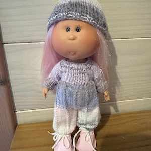 Peut inclure: Poupée faite à la main avec des cheveux roses, portant un bonnet en tricot gris et blanc, un pull assorti et un pantalon à blocs de couleurs rose, gris et blanc. La poupée a des chaussures roses avec des lacets blancs.