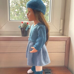 May include: A doll dressed in a blue knitted beret, sweater, and leg warmers, with a blue patterned dress and white socks. The doll is placed near a window, displaying a handcrafted ensemble.