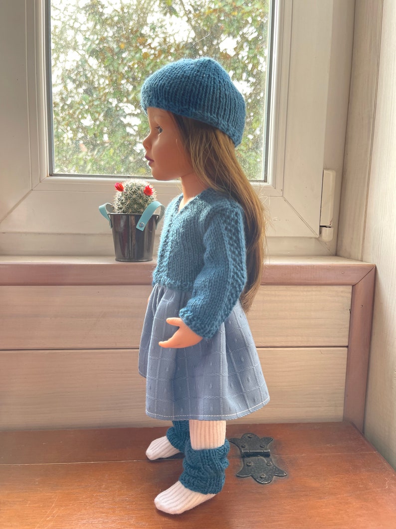 May include: A doll dressed in a blue knitted beret, sweater, and leg warmers, with a blue patterned dress and white socks. The doll is placed near a window, displaying a handcrafted ensemble.