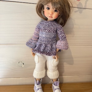 May include: A doll wearing a purple and pink knitted sweater, white knitted trousers with cuffed legs, and lavender trainers.