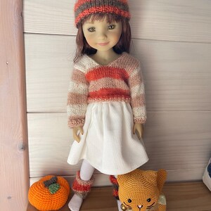 May include: A doll dressed in a striped sweater, white dress, and a matching hat. The doll has brown hair and is wearing knitted leg warmers. An orange crocheted pumpkin and a crocheted cat are placed nearby.