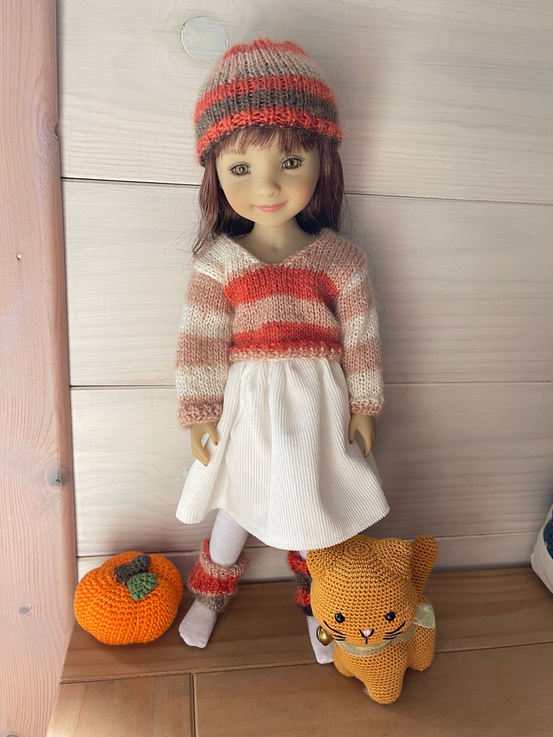 May include: A doll dressed in a striped sweater, white dress, and a matching hat. The doll has brown hair and is wearing knitted leg warmers. An orange crocheted pumpkin and a crocheted cat are placed nearby.