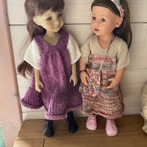 Puede incluir: Dos muñecas con vestidos de punto. La muñeca de la izquierda lleva un vestido morado con mangas blancas y botas negras. La muñeca de la derecha lleva un vestido a rayas beige y rosa con un bolso de ganchillo y zapatos rosas.