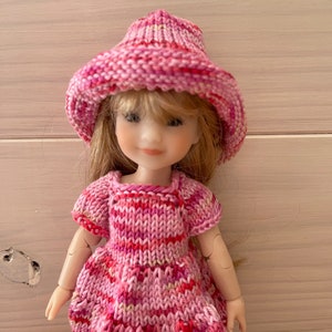 May include: A pink knitted dress and hat set for a doll. The dress has a striped pattern and the hat is a wide-brimmed sun hat.