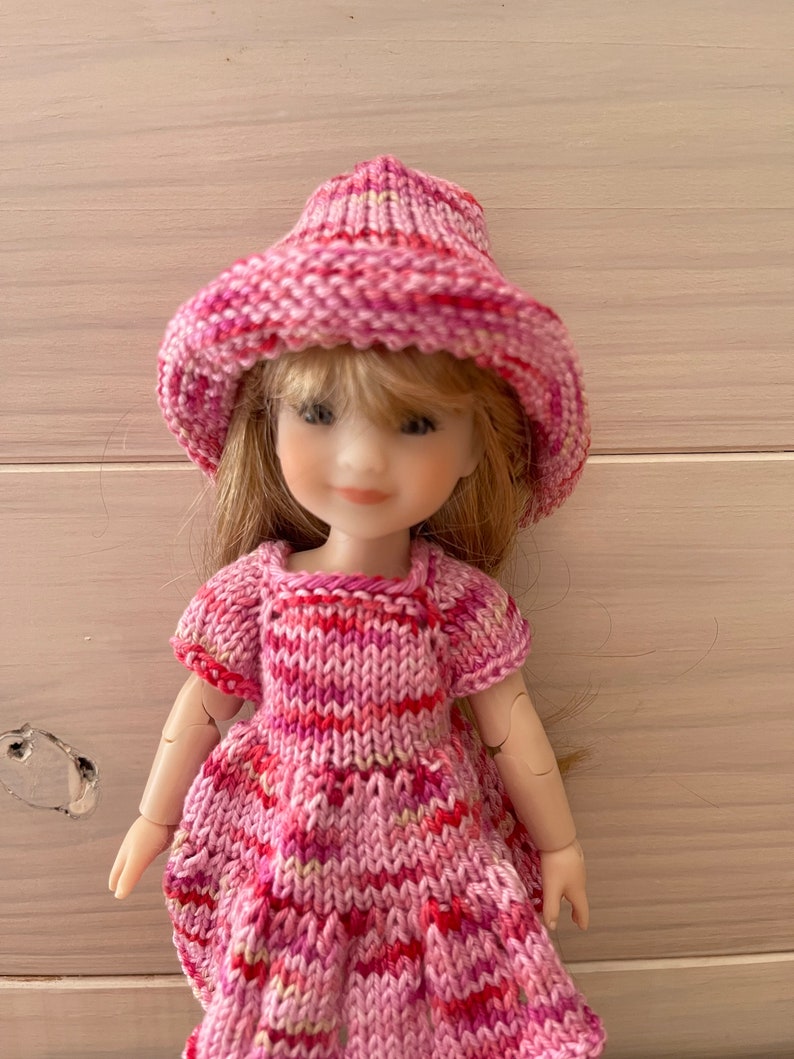 May include: A pink knitted dress and hat set for a doll. The dress has a striped pattern and the hat is a wide-brimmed sun hat.