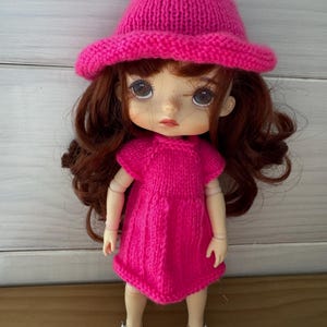 Puede incluir: Un conjunto de vestido y sombrero de punto rosa para una muñeca. La muñeca tiene el pelo castaño y lleva zapatos blancos.