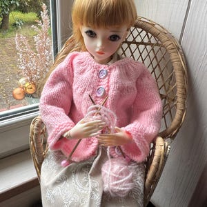 May include: A doll with blonde hair wearing a light pink knitted cardigan with three buttons, seated in a wicker chair. The doll holds two knitting needles and a ball of pink yarn, and wears a beige skirt featuring a white floral pattern.
