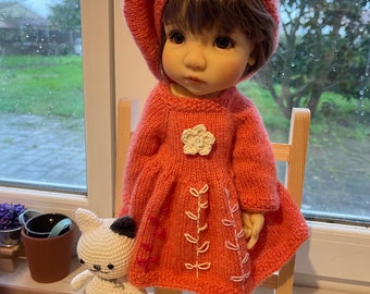 May include: A doll wearing a coral knitted dress with white embroidery, a coral beret, and white shoes. The doll is holding a white crocheted cat toy.